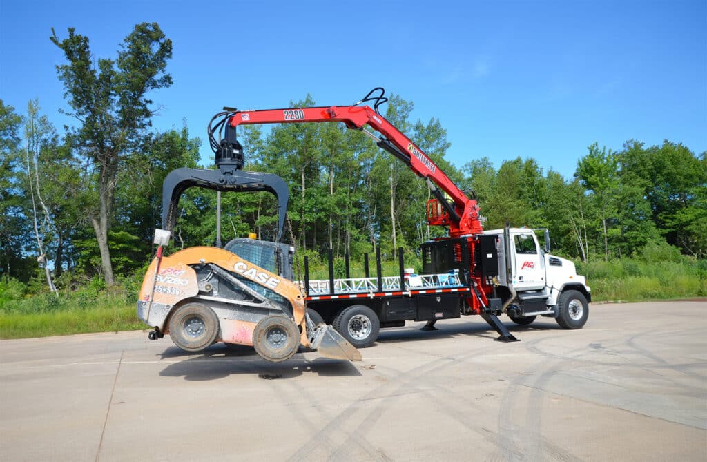 A truck equipped with a red hydraulic crane lifts a skid steer loader in a spacious outdoor area. The background features lush green trees under a clear blue sky. Tire marks are visible on the paved ground.