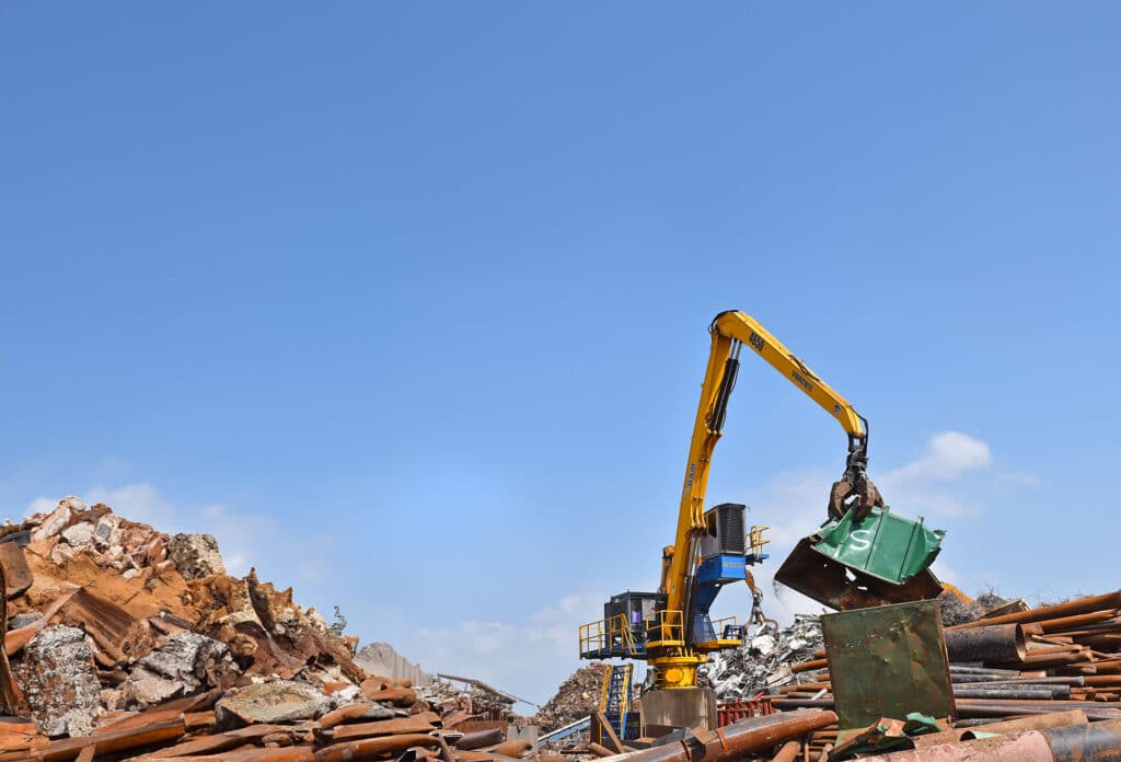 A yellow crane with a claw attachment lifts a green metal container among a pile of scrap metal under a clear blue sky.