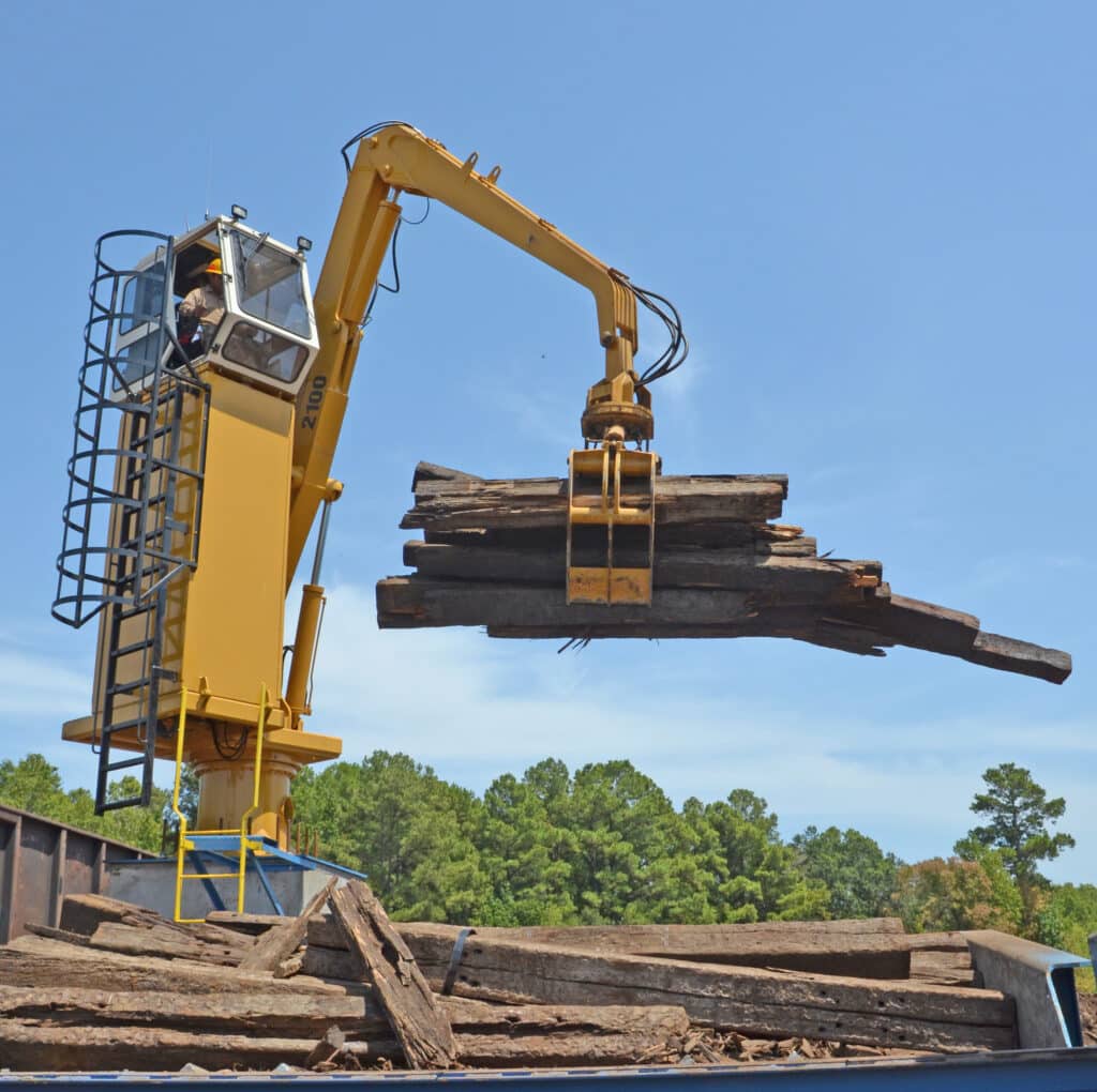 A large yellow crane with a claw attachment is lifting a load of wooden beams. The operator is inside an elevated cabin above the crane arm. The background features a clear blue sky and green trees.