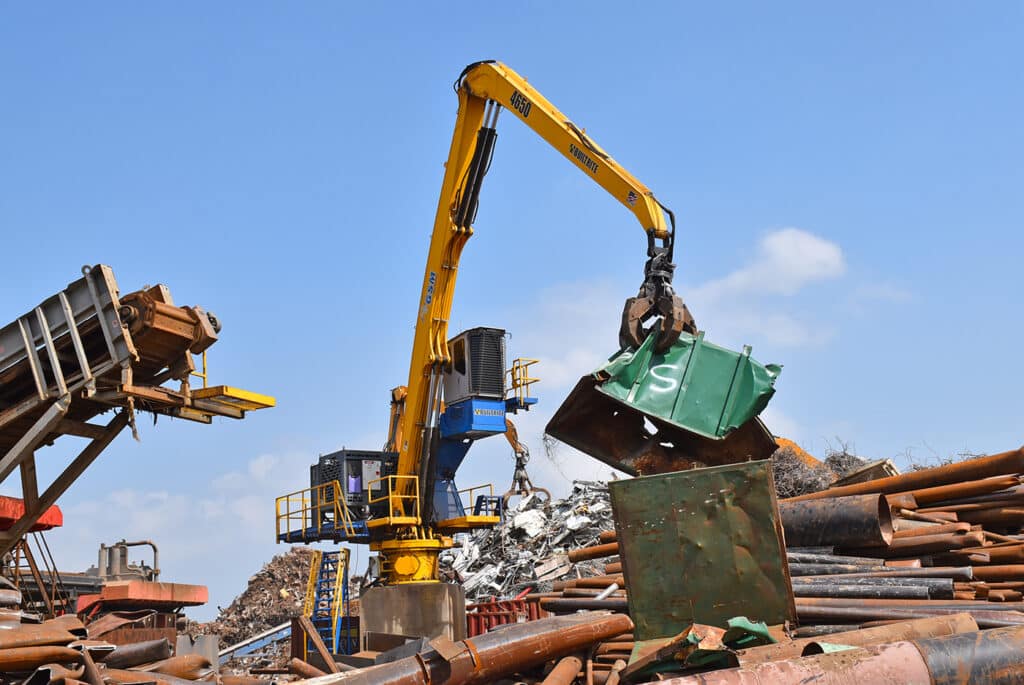 A large yellow and blue crane is lifting metal objects at a construction or industrial site. Pipes and various structures are scattered in the background under a blue sky with clouds.