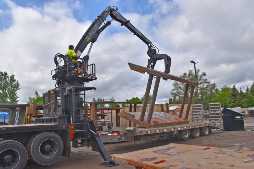 A worker in a neon yellow shirt operates a crane attached to a truck to lift large wooden beams onto a flatbed trailer in an outdoor industrial area. The sky is partly cloudy and the area is surrounded by trees and various industrial materials.