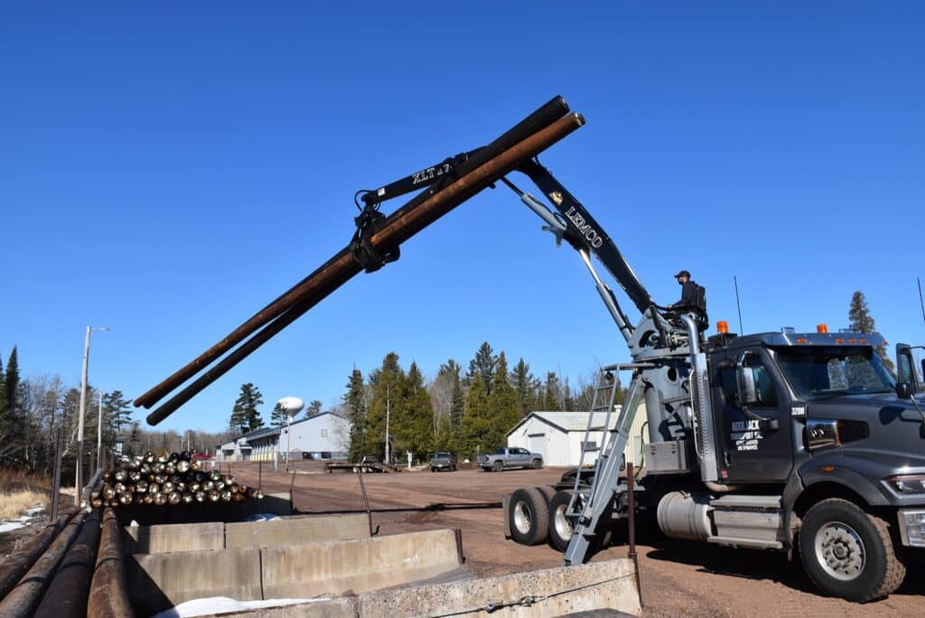 A heavy-duty truck equipped with a hydraulic crane is lifting large metal pipes at a construction site. The area is surrounded by trees and a few industrial buildings under a clear blue sky. The operator is standing on the truck controlling the crane.