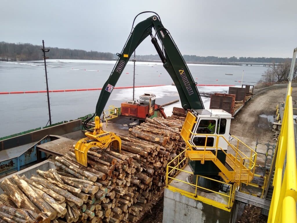 A large, green hydraulic log loader with a yellow grapple attachment is seen loading or unloading logs from a barge by a river. The area is industrial, with logs stacked on the barge and machinery in the background. The river is partially frozen.