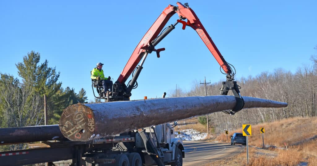 A truck-mounted crane loads a large wooden utility pole onto the back of a flatbed truck on the side of a rural road. The blue sky and bare trees indicate it's a clear, chilly day.