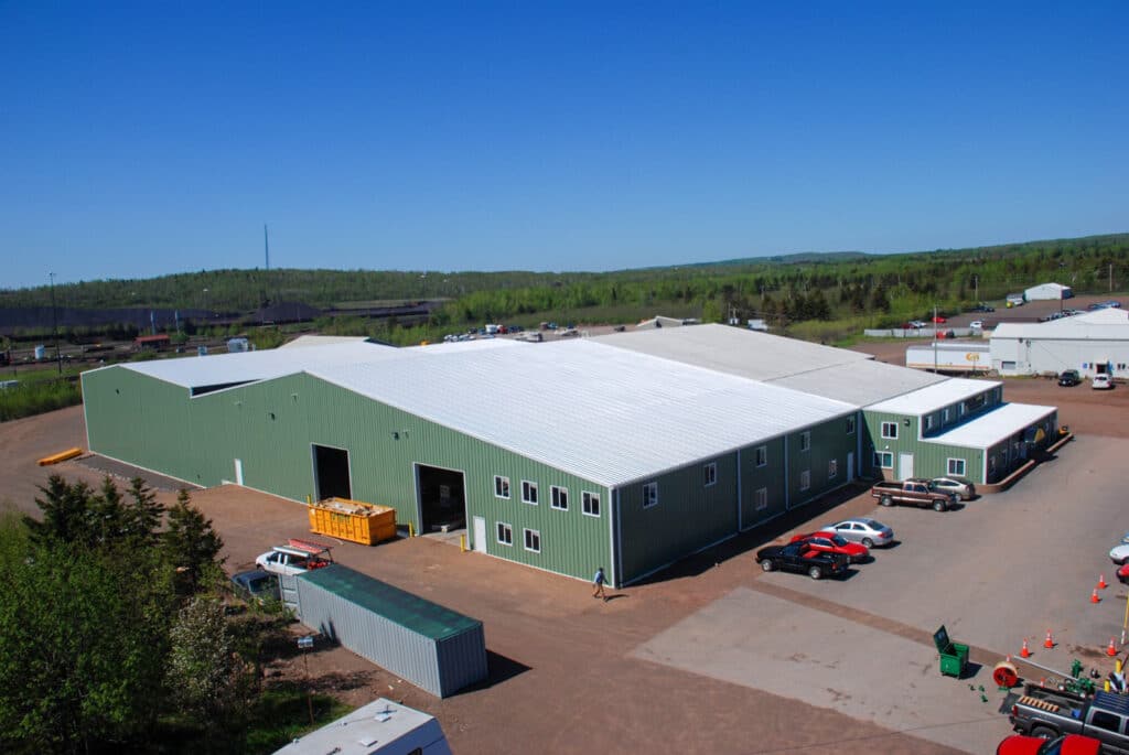 A large green industrial building with a white roof is situated in a spacious outdoor area. Several cars are parked in the lot, with various equipment visible around the building. The background consists of green fields and trees under a clear blue sky.