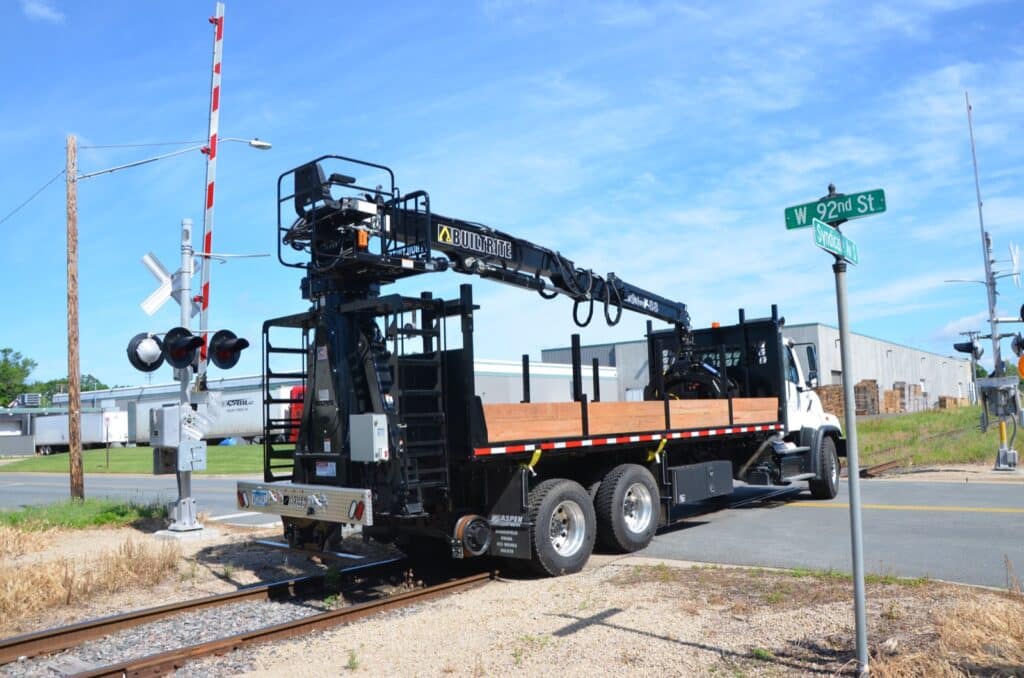 A large white utility truck with an extendable crane is stopped near a railroad crossing. There's a street sign nearby indicating "W 92nd St." and a railway signal and control box are visible in the background.