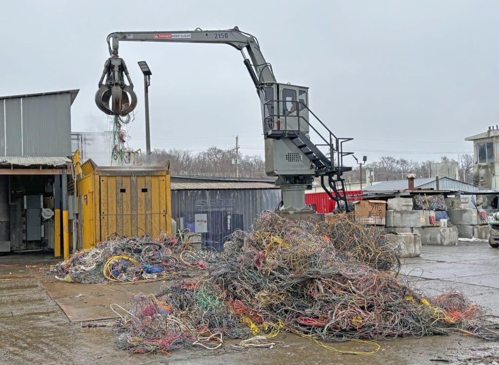 A large metal claw attached to a crane sorts through a massive pile of tangled, multicolored wires and cables at a scrapyard on a dreary, overcast day. Industrial buildings and various debris are visible in the background.