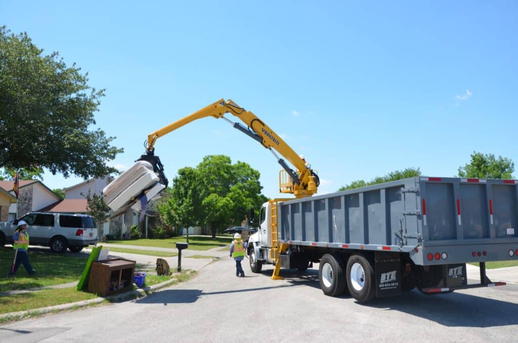 A large, yellow hydraulic crane attached to a truck lifts a residential trash container on a sunny street. Two sanitation workers in high-visibility vests stand nearby, operating the equipment. Trees and houses line the background.