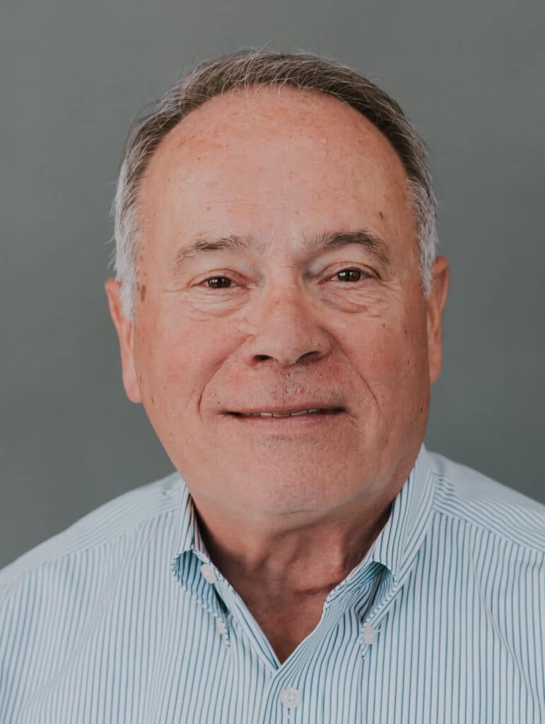 A smiling older man with short gray hair and wearing a light blue, striped button-up shirt stands in front of a plain gray background.