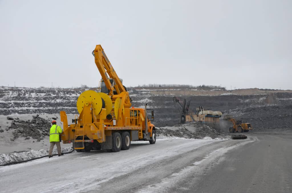 A worker in a high-visibility jacket walks towards a large yellow maintenance truck on a gravel road at an open-pit mine. Heavy machinery is operating in the background amid rocky terrain and a gray, overcast sky.