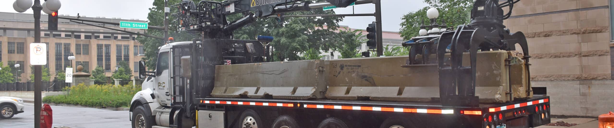 A large utility truck equipped with a crane arm is parked on a wet city street near a construction zone. Concrete barriers and orange traffic barrels surround the truck. Trees and buildings are visible in the background.