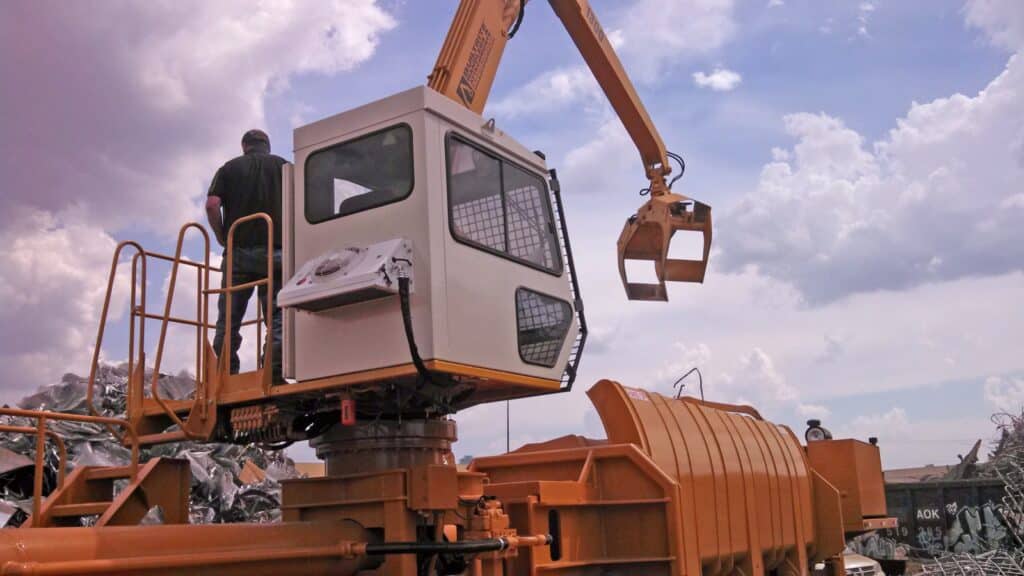 A man operates a large industrial vehicle equipped with a mechanical arm and a heavy-duty gripper. He is standing on an elevated platform next to the vehicle, amidst piles of scrap metal under a partly cloudy sky.