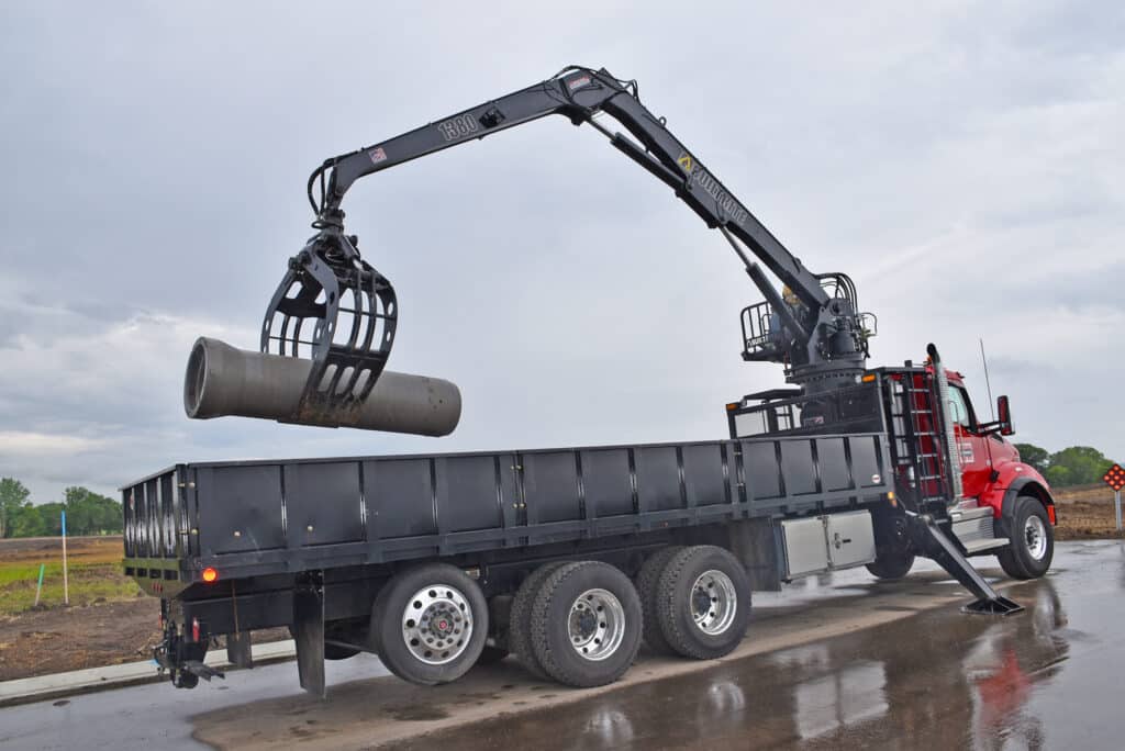 A red truck with a black flatbed is parked on a wet road. It has a crane attached to it, lifting a large concrete pipe with its claw. The sky is overcast, and there is construction work visible in the background.